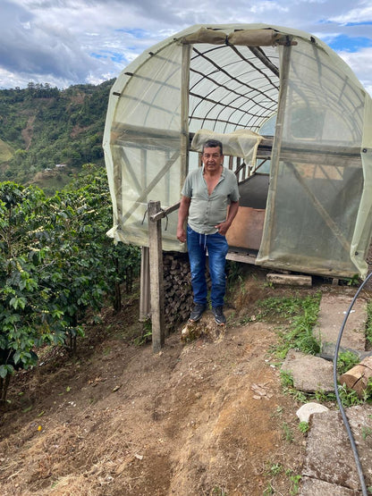 Don Franco Mutis Diaz stands in front of a small greenhouse or hoop house made of translucent plastic over a wooden frame, with coffee trees in the background.